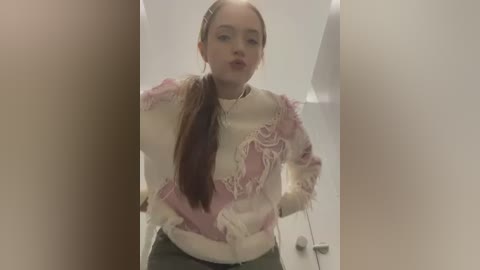 Media: Video of a young woman with fair skin, brown hair, wearing a frayed pink and white sweater, standing in a dimly lit bathroom with white walls and a closed door.