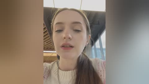 Media: Video of a young Caucasian woman with light skin and straight brown hair tied in a ponytail, wearing a knitted beige sweater, sitting inside a car with a light-colored interior.