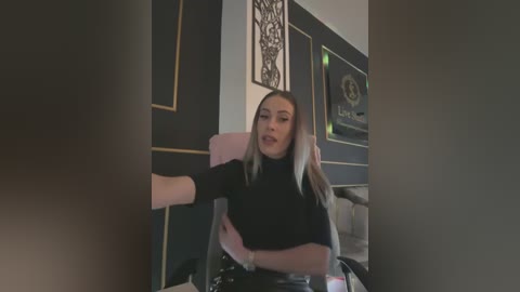 Media: Video of a young woman with light skin, straightened blonde hair, wearing a black turtleneck sweater, seated in a pink chair with a white armrest, in a modern room with dark walls and framed artwork.