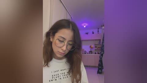 Media: Video of a young woman with long brown hair and glasses, wearing a white T-shirt with a motivational quote, standing in a brightly lit, modern kitchen with purple lighting.