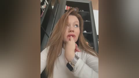 Media: Video of a Caucasian woman with long, light brown hair, wearing a white top, and a silver watch. She's standing in an office with a black filing cabinet and a whiteboard in the background. She's biting her index finger in a contemplative pose.