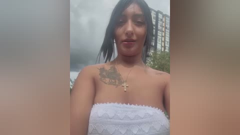 Media: Video of a young, slender woman with dark hair and light skin, wearing a white bandeau top, standing outdoors near a cloudy sky and urban buildings.