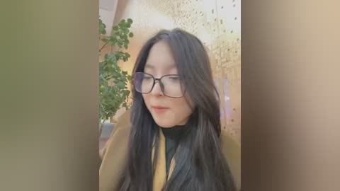 Media: Video of an Asian woman with long black hair, wearing black-rimmed glasses, a mustard-yellow top, and a black vest, smiling softly, against a beige, perforated wall background with a green potted plant to the left.