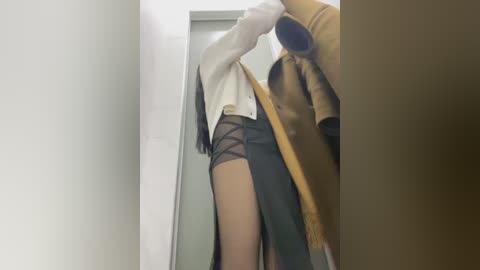 Media: Video of a slender, light-skinned woman with a medium build, wearing a white jacket and sheer black shorts, holding a beige coat and standing in a narrow, dimly-lit hallway.