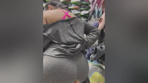 Media: Video of a woman with dark hair in a grey, long-sleeved shirt and pink bra, bending over in a crowded, dimly lit room filled with clothing racks and people.