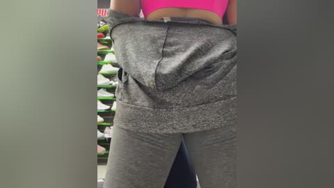 Media: Video of a woman in a gray athletic outfit, holding up her pants, showing a bright pink sports bra, against a backdrop of colorful shoe shelves.