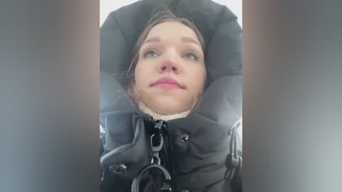 Video of a woman with fair skin and light brown hair, wearing a black puffy jacket and a dog collar, looking up with a neutral expression.