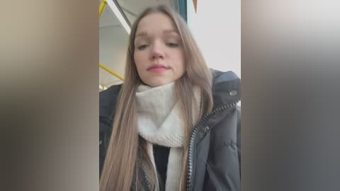 Media: Video of a young Caucasian woman with long, straight brown hair, wearing a black puffer jacket and white turtleneck, standing inside a bus with yellow seats visible in the background.