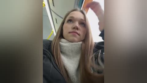 Media: A video of a young Caucasian woman with long, straight, light brown hair, wearing a black jacket and beige turtleneck sweater, sitting in a train carriage, looking contemplatively through the window.