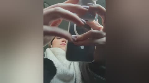 Media: A video shows a woman's hands holding a smartphone, capturing a selfie with blurred facial features. She wears a white sweater, seated in a dark car.