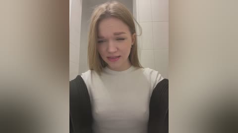 Video of a young, light-skinned, blonde woman in a bathroom stall, wearing a black cardigan over a white ribbed top, looking down with a slight smile.