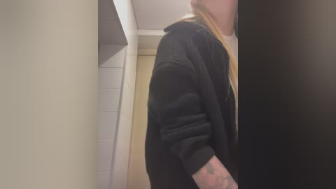 Video of a young woman with long blonde hair, wearing a black cardigan over a white shirt, leaning against a tiled wall in a dimly lit bathroom.