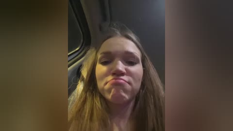 Media: Video of a young woman with long, straight brown hair, puckering her lips in a pout, taken from inside a car. The background shows a dark, windowless interior.