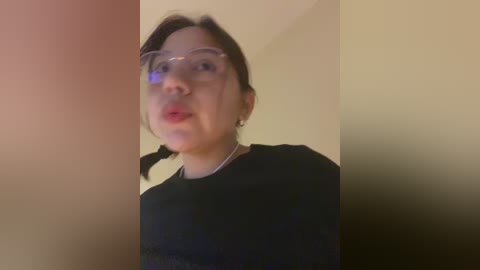 Media: Video of a young East Asian woman with fair skin, dark hair, and clear glasses, wearing a black shirt. She is blowing a bubble with her red lips. The background is blurred.