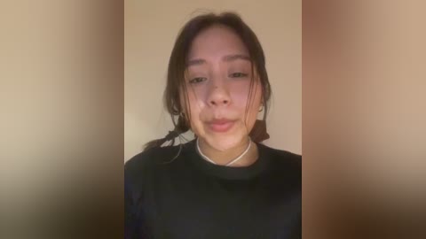 Media: Video of a young Asian woman with long dark hair, wearing a black shirt, against a beige wall. She has a neutral expression.