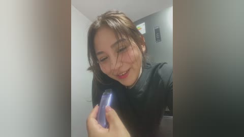 Media: A video of a smiling East Asian woman with long dark hair, wearing a black top, holding a blue phone, in a dimly lit room with white walls and a framed picture.