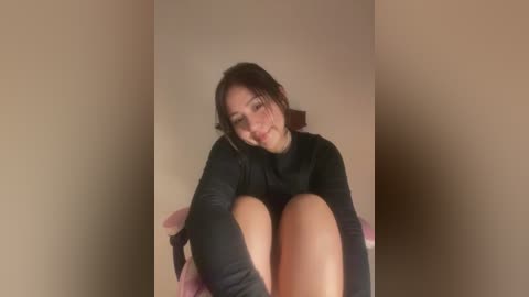 Media: Video of a young woman with light brown skin, dark hair, and wearing a black turtleneck, sitting on a pink chair against a beige wall. She has a relaxed, slightly playful expression.