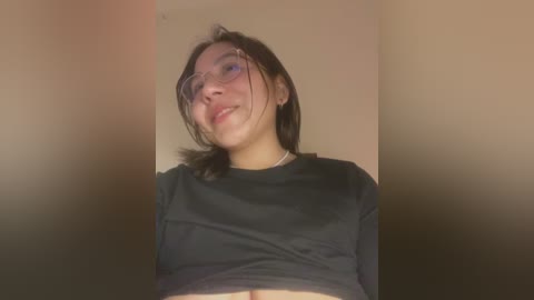 Media: Video of a young Asian woman with fair skin, short dark hair, and glasses, smiling slightly. She wears a black crop top, revealing her midriff, against a beige background.