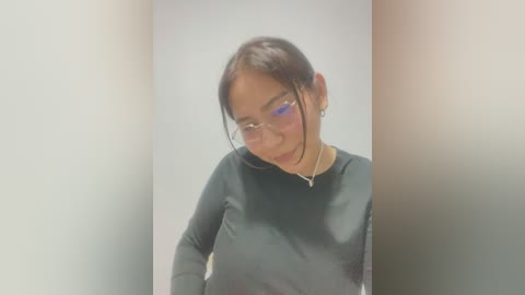 Media: Video of a young woman with dark hair and light skin, wearing glasses and a black long-sleeve top. She has a neutral expression. The background is a blurred gradient of light and dark colors.