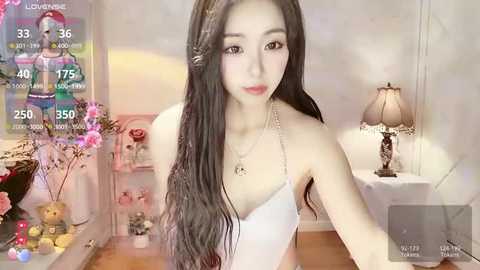 Media: A video of an Asian woman with long black hair, wearing a white spaghetti-strap top, standing in a softly lit room with pink cherry blossoms, a lamp, and a digital screen displaying stats.