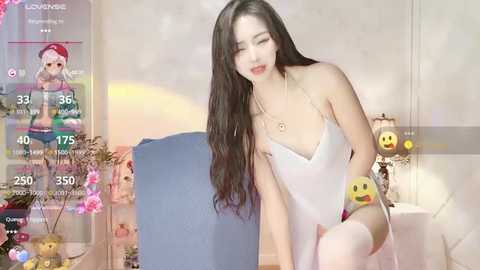 Media: Video of a fair-skinned, long-haired Asian woman in a white lingerie set, sitting seductively on a blue couch in a room with a pink wall. The scene is overlaid with social media app elements.