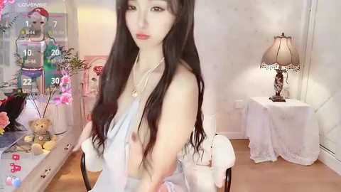 Media: A video of a young Asian woman with long black hair, fair skin, and small breasts, wearing a sheer white top, standing in a cozy, softly lit room with floral decorations, a table lamp, and a teddy bear.