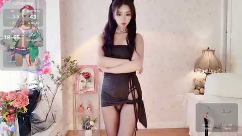 Media: Video of an East Asian woman with long black hair, wearing black lingerie and a choker, standing in a softly lit room with floral decor and a lamp.