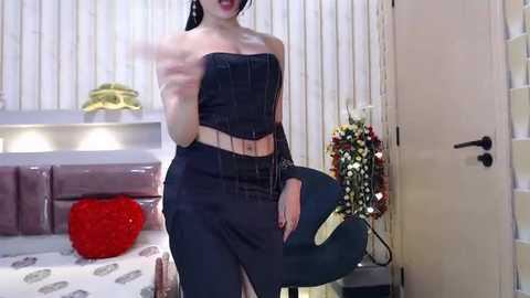 Media: Video of a slender woman with fair skin and long dark hair, wearing a black strapless corset and high-waisted pants, standing in a modern bedroom with a heart-shaped pillow on a bed and floral arrangement near a door.