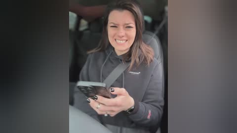 Media: Video of a Caucasian woman with light skin and long brown hair, smiling while holding a smartphone, wearing a black hoodie and gray seatbelt, seated in a car.