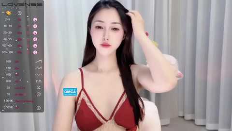 Media: A video of a young East Asian woman with long black hair, fair skin, and red lipstick, wearing a revealing red lingerie top with beige lace trim. She's seated indoors with white curtains in the background.