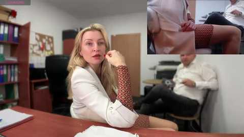 Media: Video of a blonde woman in a white blouse, red-patterned sleeves, sitting at a desk, looking thoughtful, in an office with bookshelves and a man in a white shirt and black pants, seated.