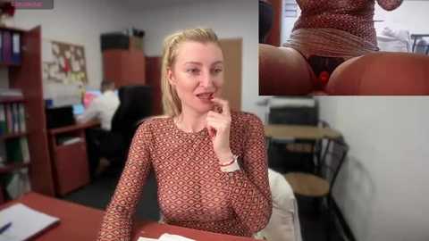 Media: Video of a blonde, fair-skinned woman with medium-sized breasts, wearing a patterned long-sleeve top, seated at a desk in an office, biting her finger suggestively, with a close-up inset of her exposed crotch.