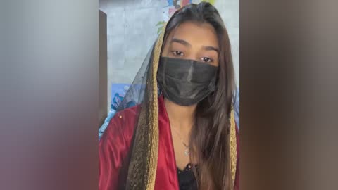 Media: Video of a young woman with medium-brown skin and long black hair, wearing a red kurta, black face mask, and gold headscarf. Background shows a blurred, colorful wall and door.