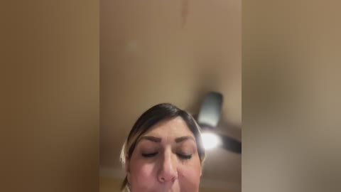 Media: A video of a woman with closed eyes, light skin, and short dark hair, taking a selfie in a dimly lit room with beige walls and a lamp in the background.