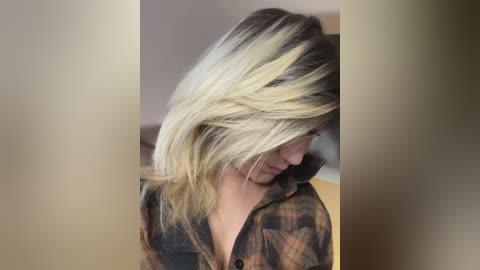 Media: Video of a fair-skinned woman with shoulder-length, blonde and dark brown ombre hair, wearing a brown and black plaid shirt, looking downward. Background is blurred with a neutral color palette.