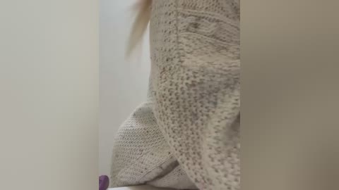 Media: Video of a close-up view of a person wearing a light beige, textured, crocheted sweater, with a subtle purple garment partially visible in the background. The image is slightly blurred, focusing on the textures and colors of the clothing.