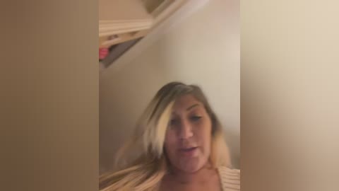 Media: A blurry video of a young woman with long blonde hair, fair skin, and a neutral expression, standing in a dimly lit room with beige walls and a visible white doorframe in the background.