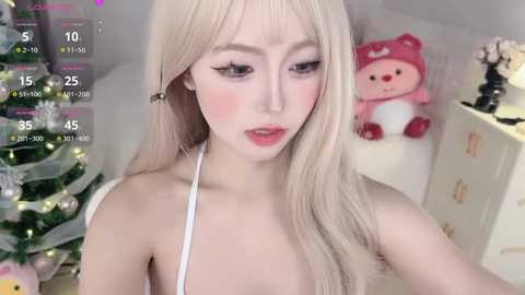 Media: Video of an East Asian woman with long, platinum blonde hair, fair skin, and pink lipstick, wearing a white top, standing in a cozy room with a Christmas tree and a pink plush toy in the background.