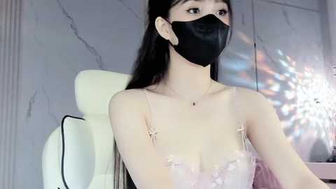 Media: Video of an East Asian woman with long black hair, fair skin, wearing a black face mask, delicate pink lace dress, and sitting in a white chair against a marble wall background.