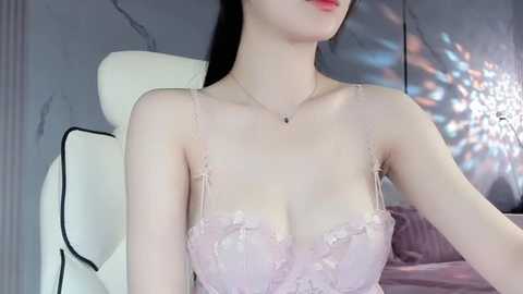 Media: Video of a slender, pale-skinned Asian woman with long black hair, wearing a light pink lace bra. She sits on a white leather chair in a dimly lit room with a marble-patterned wall.