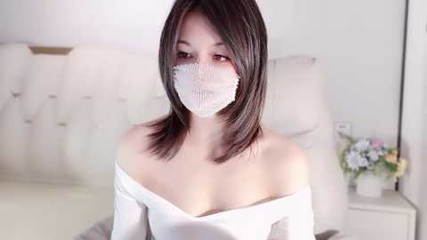 Media: Video of an Asian woman with shoulder-length black hair, wearing a white off-shoulder top, a white mask, and seated in a modern, minimalistic bedroom with white furniture and a floral arrangement on a nightstand.