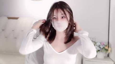 Media: Video of an Asian woman with light skin, straight dark brown hair, and a white face mask, adjusting her hair in a white long-sleeved top. Background features a white bed with a tufted headboard, a white nightstand with a vase of flowers, and soft lighting.