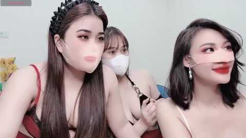 Media: Video of three East Asian women with fair skin, wearing face masks, black headbands, and red lipstick, sitting closely together, smiling.