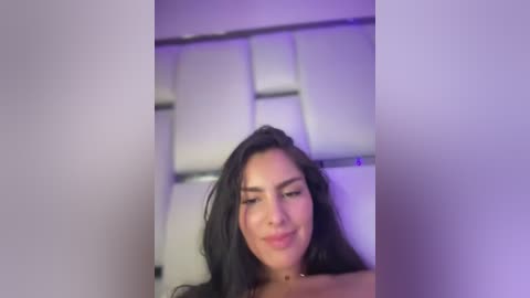 Media: Video of a young Latina woman with long black hair, wearing a light-colored top, smiling. She is indoors with a modern, minimalist background featuring white, geometrically shaped wall panels and purple lighting.