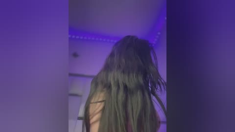 Media: Video of a woman with long, wet, dark hair, standing in a dimly lit room with purple lighting, wearing a sheer top, facing away from the camera.