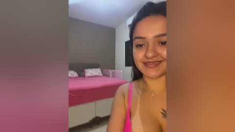 Media: A video shows a smiling Latina woman with long black hair, wearing a pink spaghetti strap top and sitting in a modern bedroom with a pink bed and beige walls.