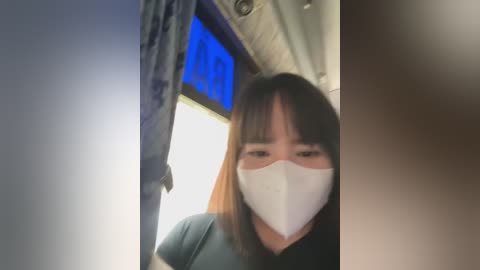 Media: A video of a young woman with straight, shoulder-length brown hair and light skin, wearing a white surgical mask, standing in a dimly lit room with a blue curtain and wooden ceiling.