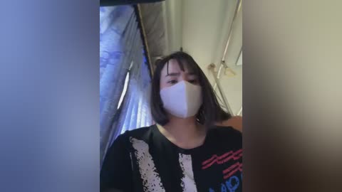 Video of a young person with a white mask, black shirt with red and blue design, and straight brown hair, standing in a dimly lit room with plastic sheeting.