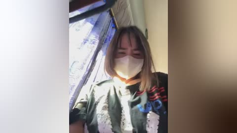 Media: A video shows a person wearing a face mask and a dark, patterned shirt, standing near a window with a blue curtain. The image is slightly out of focus, with a soft, warm lighting effect.