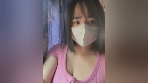 Media: Video of an Asian woman with short black hair, wearing a pink shirt and white mask, standing in a dimly lit room with a floral curtain in the background.